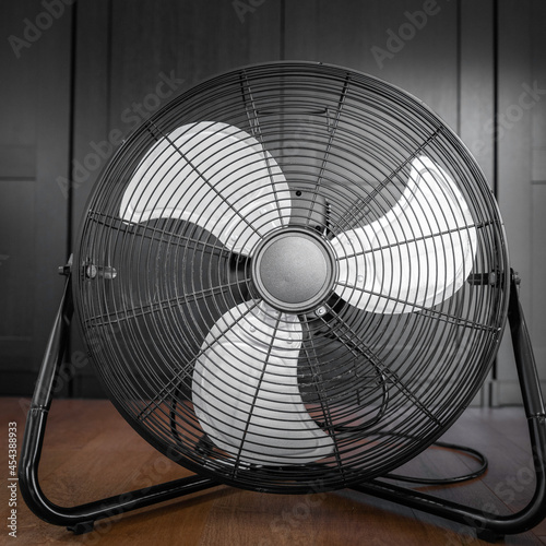 Black electric fan with three large gray metal blades. Used unplugged fan on the wooden floor in front of the black closet. Convenient and cheap summer home cooler machine.