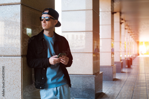 Millenial hipster guy in stylish outfit standing outdoor talking on smartphone