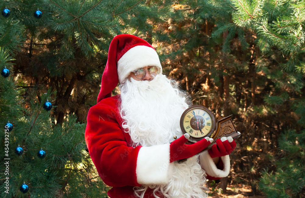 Santa Claus in a red suit against the background of green Trees of the forest in his hands holds an old wooden clock that shows 6 hours with space for text at Christmas Santa Claus in a red suit against the background of green Trees of the forest in his hands holds an old wooden clock that shows 6 hours with space for text at Christmas