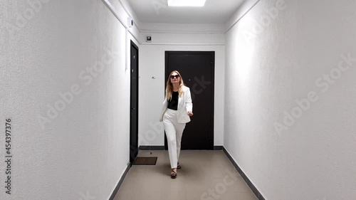 White woman in white suit confidently walks down a white corridor toward camera photo