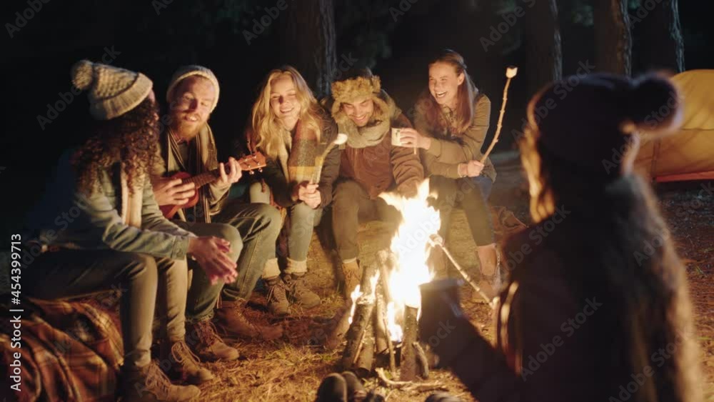 happy friends sitting by campfire young man playing ukulele singing enjoying evening together camping in forest at night happy friends sitting by campfire young man playing ukulele singing enjoying evening together camping in forest at night