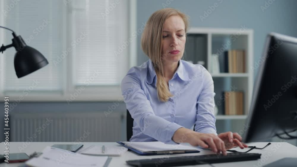 Focused mature woman in eyeglasses and formal wear writing on papers and typing on laptop while sitting at desk. Caucasian female with blond hair working at office center. Focused mature woman in eyeglasses and formal wear writing on papers and typing on laptop while sitting at desk. Caucasian female with blond hair working at office center.
