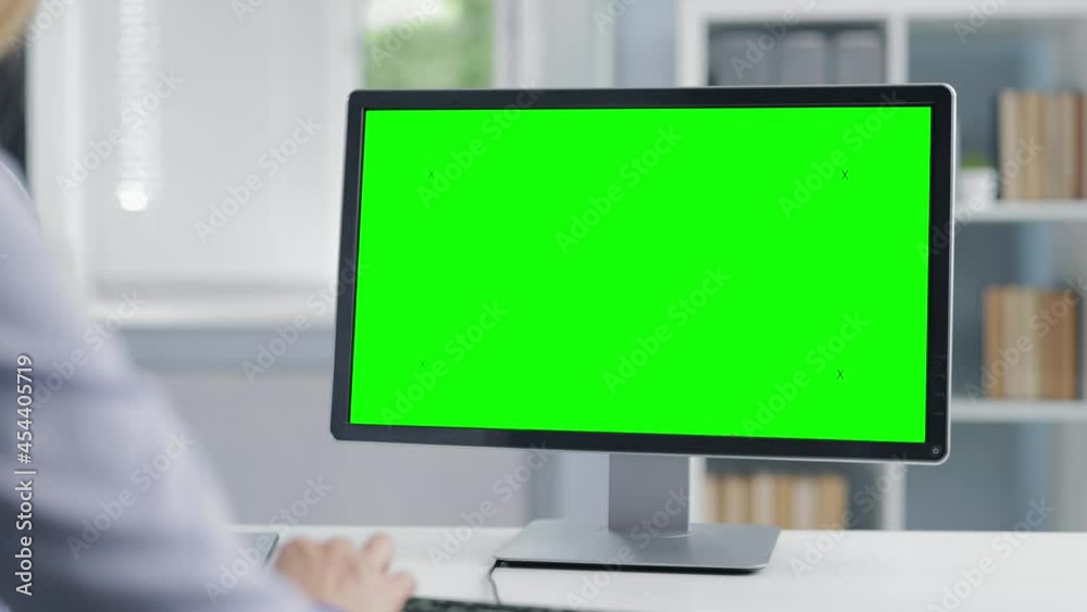 View from shoulder of woman sitting at bright office and typing keyboard. Mature lady in formal wear using computer with green empty screen. View from shoulder of woman sitting at bright office and typing keyboard. Mature lady in formal wear using computer with green empty screen.