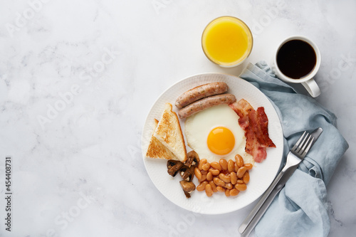 Horizontal overhead of classic morning fat english breakfast with fried egg yolks, grilled sausages, roasted bacon, kidney beans, mushrooms, toast, fresh orange juice and hot coffee. Light copy space