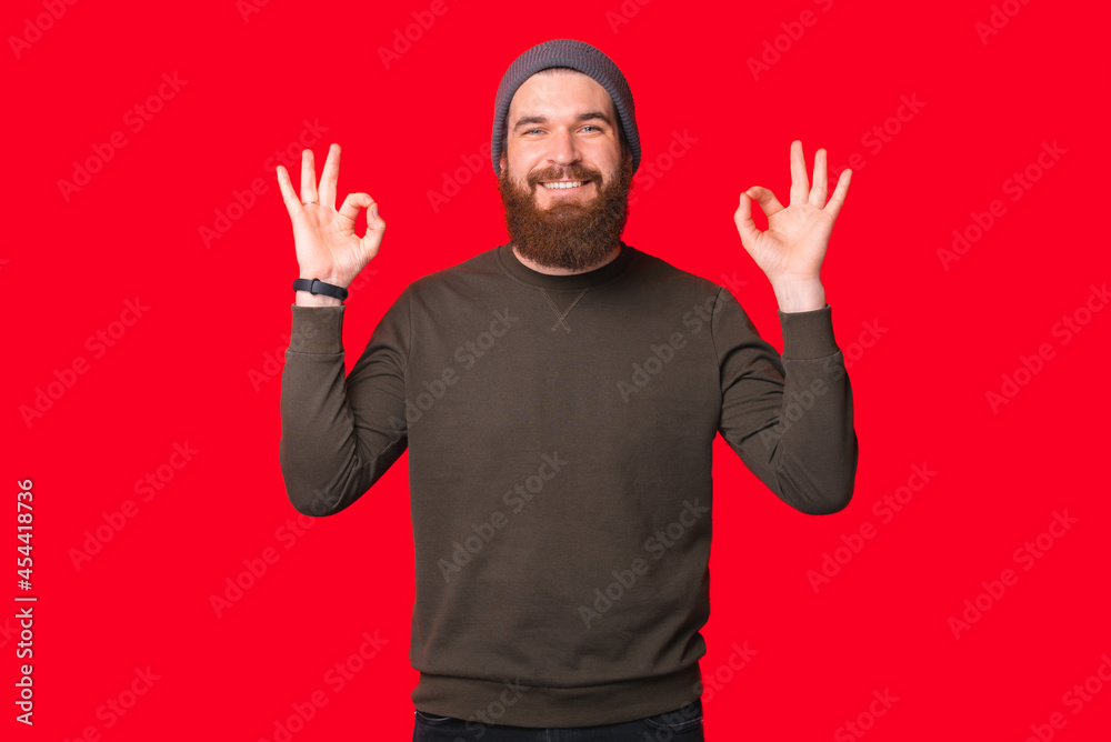 Fototapeta premium Pleased bearded man is showing ok gesture over red background while smiling at the camera.