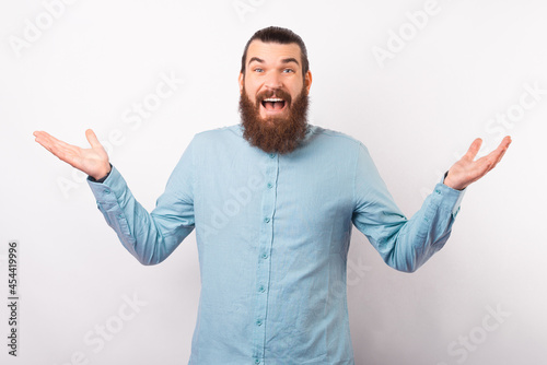 Wide smiling young bearded man shrugs over white background in a studio.