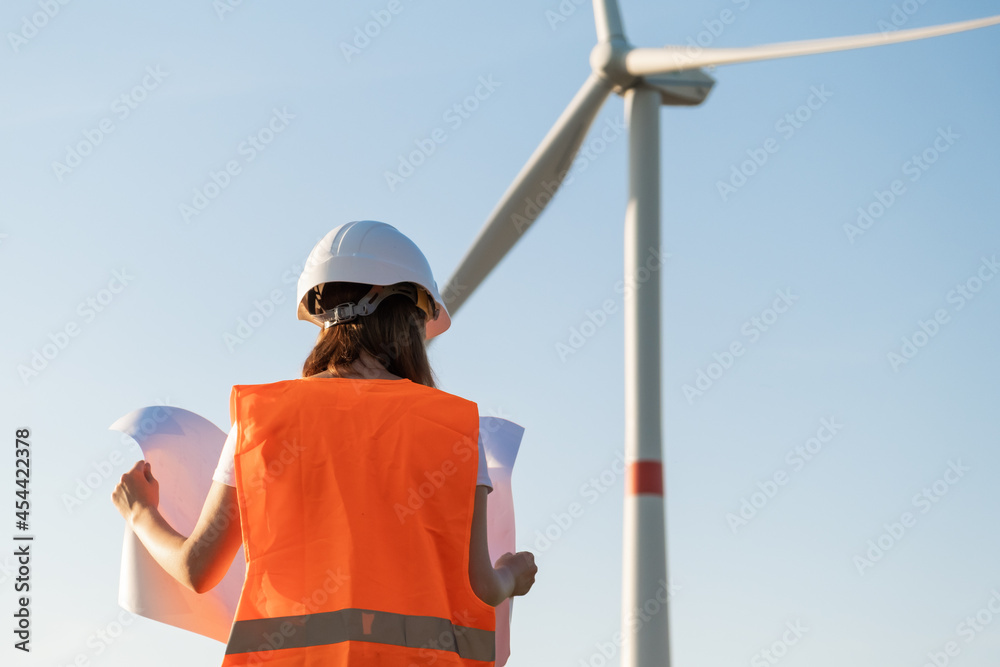Engineer in hard hat and protective vest holds a paper plan project for the construction of wind turbines in the field against the background of windmills. Generation of electricity. Alternative