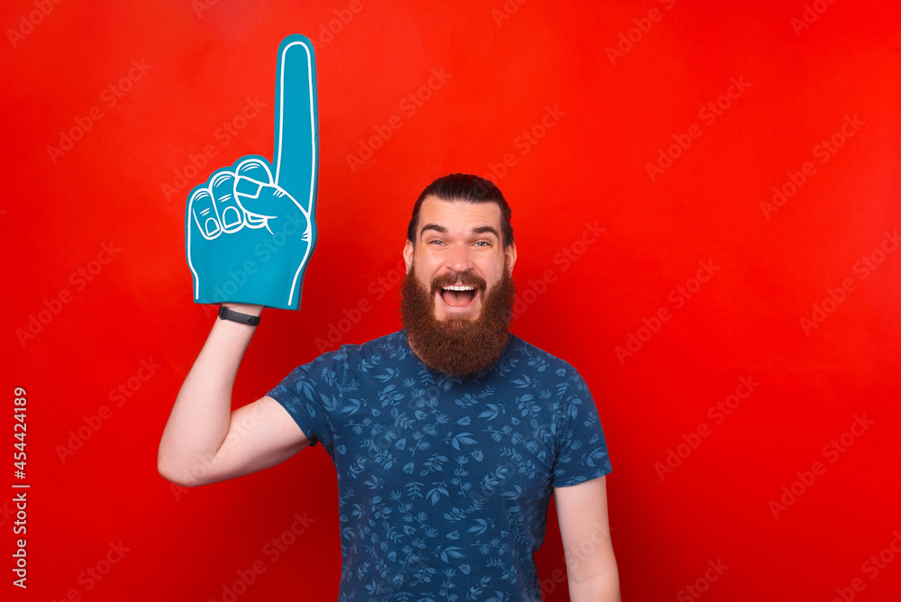 I number one fan. Excited bearded man is wearing foam glove and pointing up over red background.