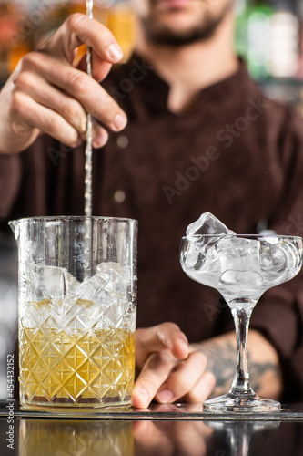 Professional bartender stirs cold cocktail with spoon. Several bottles of alcoholic drinks and glasses standing on the bar counter.