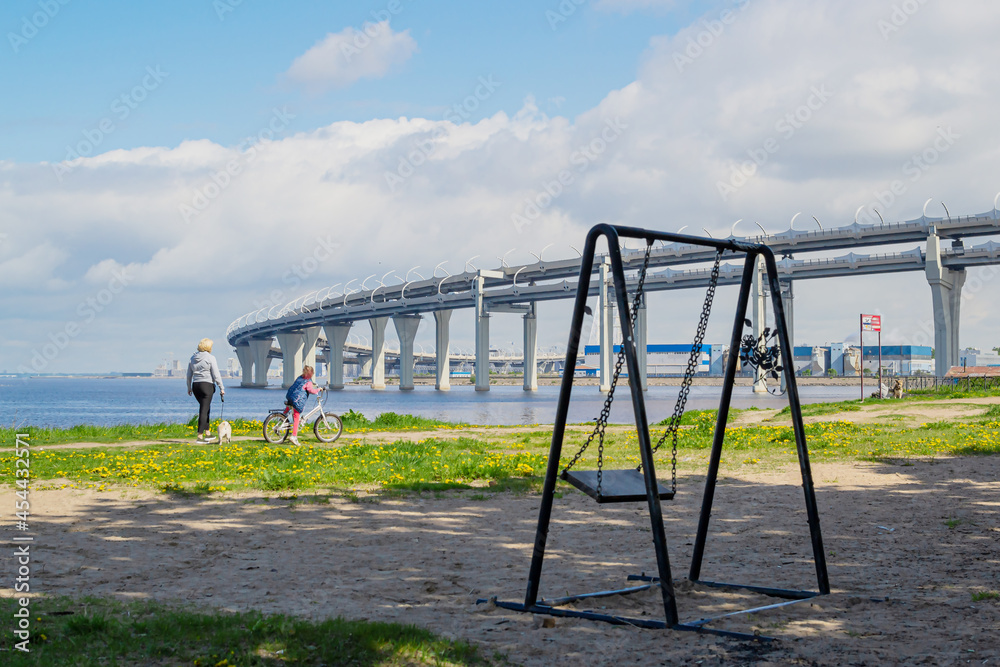 Empty swing swayed by the wind in shade of trees on the coastline with people seashore near of a big rounded motorway highway bridge, the road passing over the sea bay coast Empty swing swayed by the wind in shade of trees on the coastline with people seashore near of a big rounded motorway highway bridge, the road passing over the sea bay coast