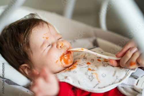 First meal problems concept small caucasian baby five months old refusing to eat spitting organic carrot mash puree while lying in a swing and hand of unknown mother holding the spoon while feeding