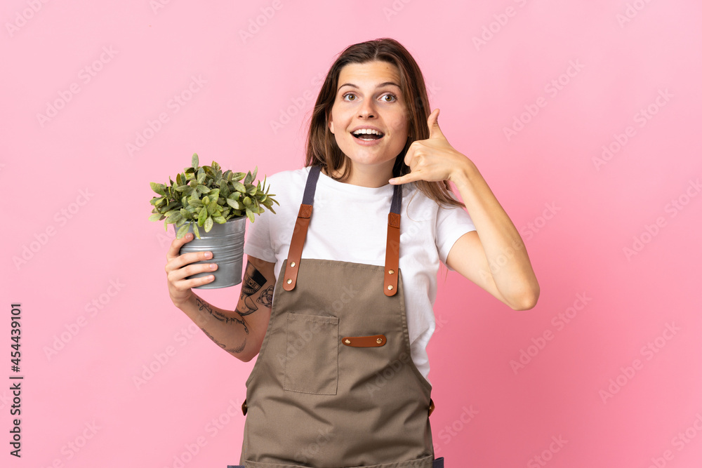 Gardener woman holding a plant isolated on pink background making phone gesture. Call me back sign Gardener woman holding a plant isolated on pink background making phone gesture. Call me back sign