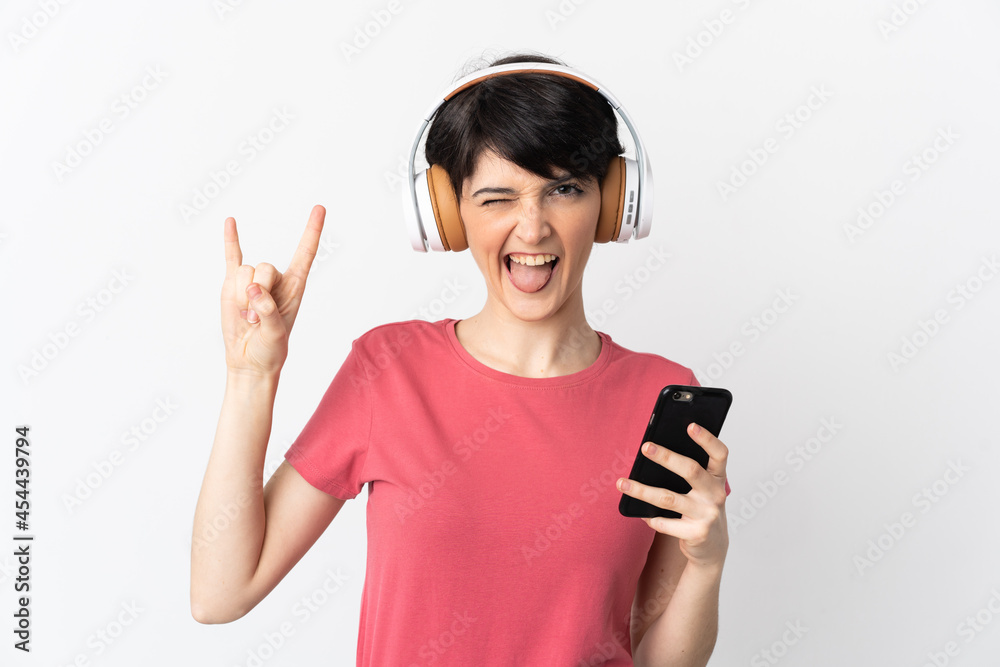 Woman with short hair isolated on white background listening music with a mobile making rock gesture