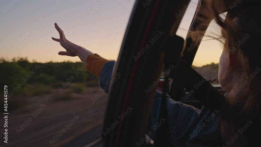 woman in car holding hand out window feeling wind blowing through fingers driving in countryside on road trip travelling for summer vacation enjoying freedom on the road at sunrise