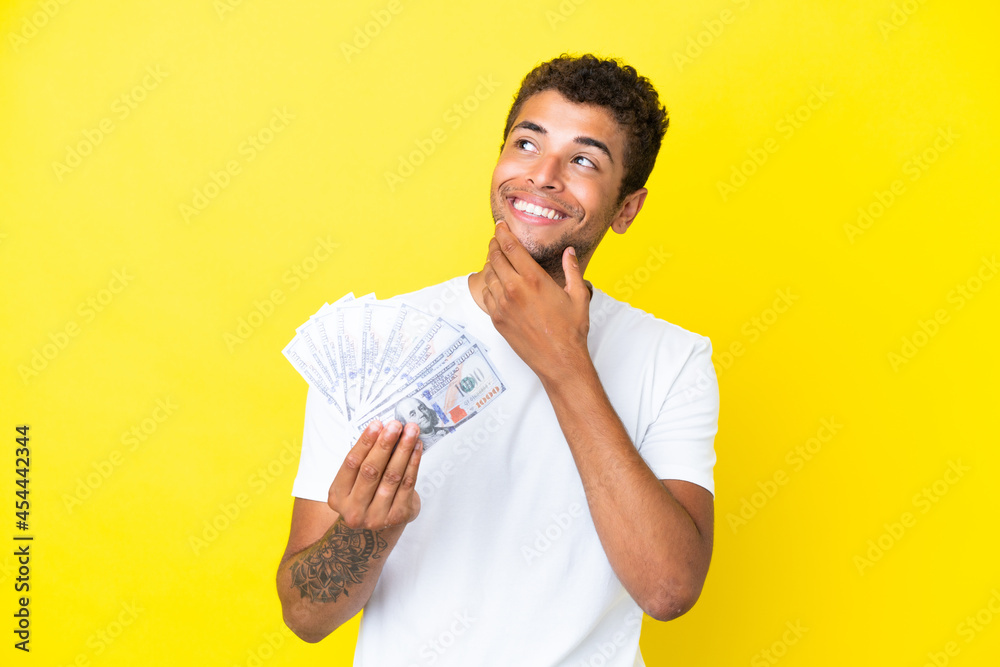 Young brazilian man taking a lot of money isolated on yellow background looking up while smiling Young brazilian man taking a lot of money isolated on yellow background looking up while smiling