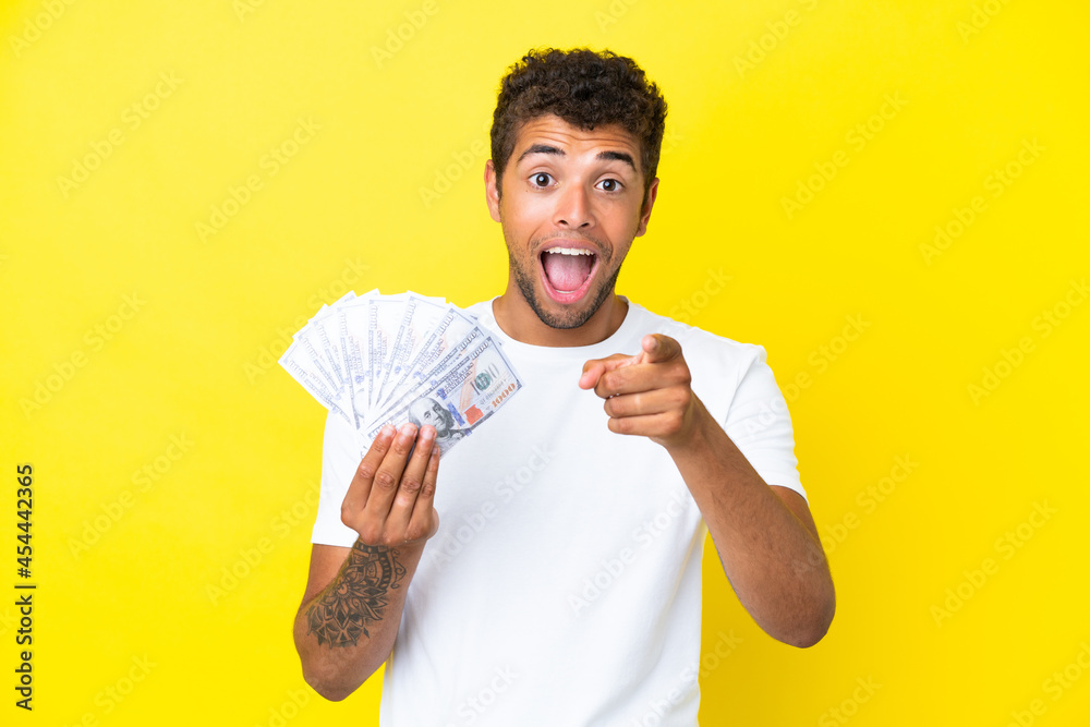 Young brazilian man taking a lot of money isolated on yellow background surprised and pointing front Young brazilian man taking a lot of money isolated on yellow background surprised and pointing front