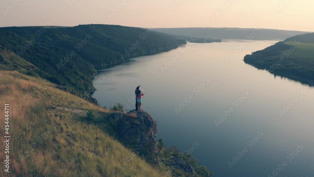 The man and woman standing on rocky mountain edge against the river The man and woman standing on rocky mountain edge against the river