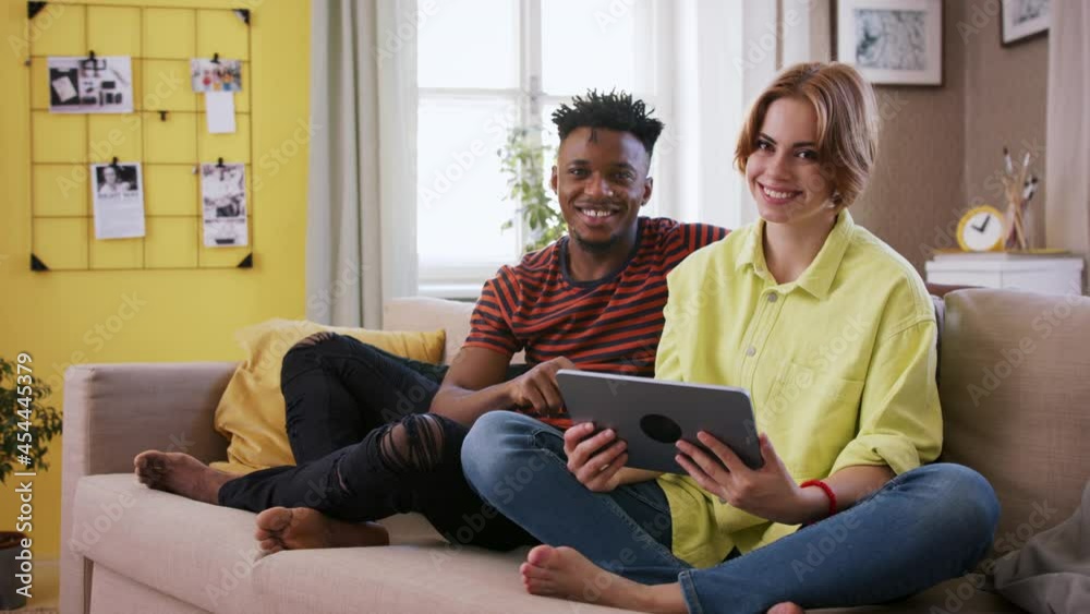 Cheerful teenagers sitting on sofa, using tablet, talking and looking at camera. Social networks concept. Cheerful teenagers sitting on sofa, using tablet, talking and looking at camera. Social networks concept.