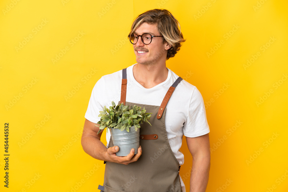 Gardener blonde man holding a plant isolated on yellow background looking to the side and smiling Gardener blonde man holding a plant isolated on yellow background looking to the side and smiling