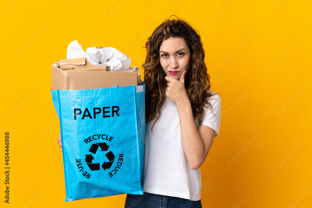 Young woman holding a recycling bag full of paper to recycle isolated on yellow background thinking Young woman holding a recycling bag full of paper to recycle isolated on yellow background thinking