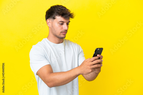 Young caucasian handsome man isolated on yellow background sending a message or email with the mobile © luismolinero