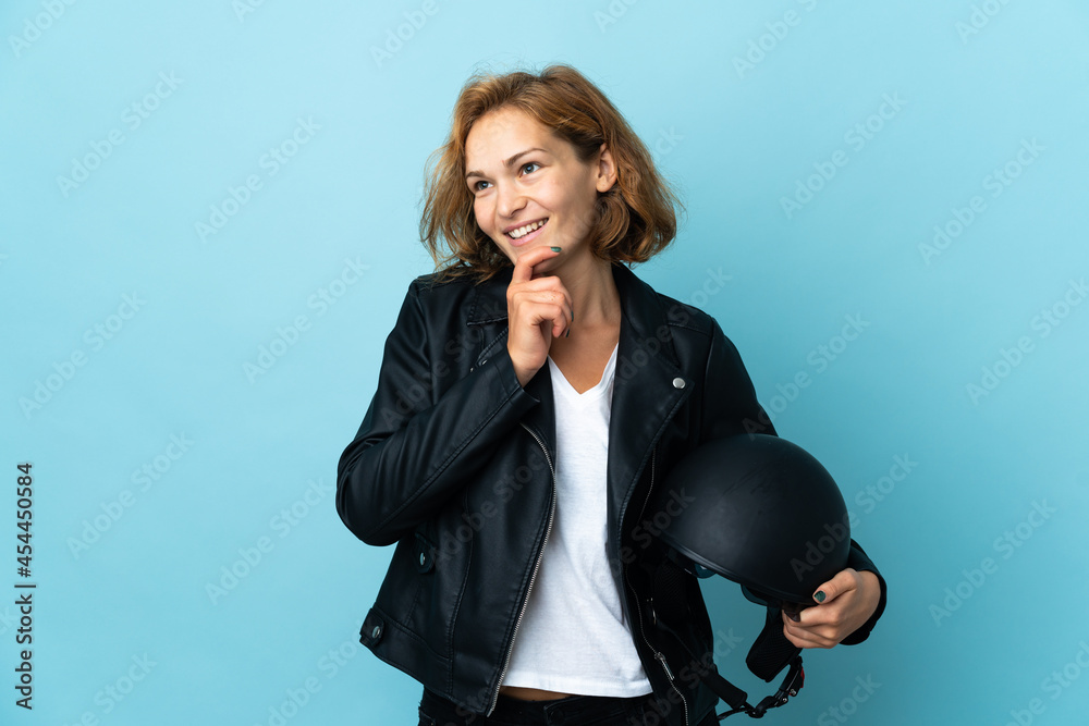 Georgian girl holding a motorcycle helmet isolated on blue background looking up while smiling Georgian girl holding a motorcycle helmet isolated on blue background looking up while smiling