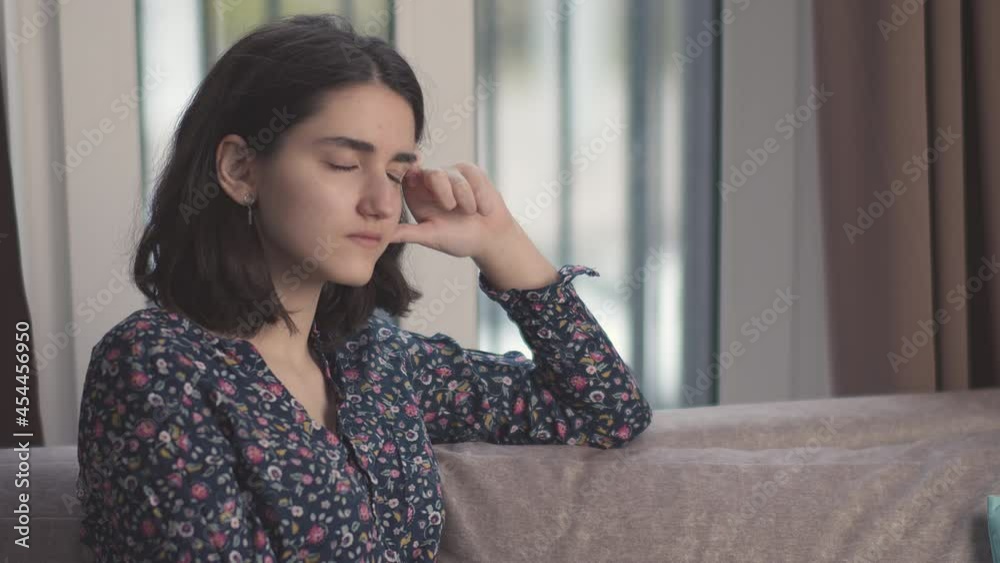 Sad teenage girl sitting on the couch with her head resting on her hand, window background