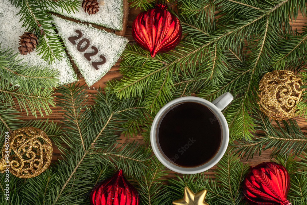 Cup of coffee and a white festival cake decorated with the number 2022 made of chocolate on the table with coniferous branches and Christmas toys. Concept of the New Year