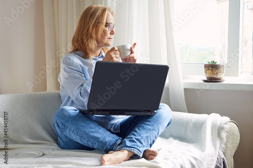 Senior woman drinking a cup of coffee during a break from remote work at a laptop at home