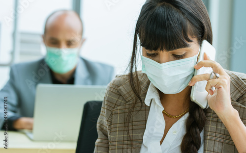 Smart Caucasian businesswoman wearing face mask protect virus, new normal, social distancing, talking on mobile phone with customer, working with intention in office with blur background of colleague.