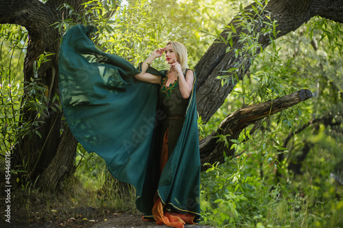 A blonde woman in a green druid cloak and in a fantasy dress with leaves. A sorceress girl in medieval clothes stands in the forest near a reservoir. photo