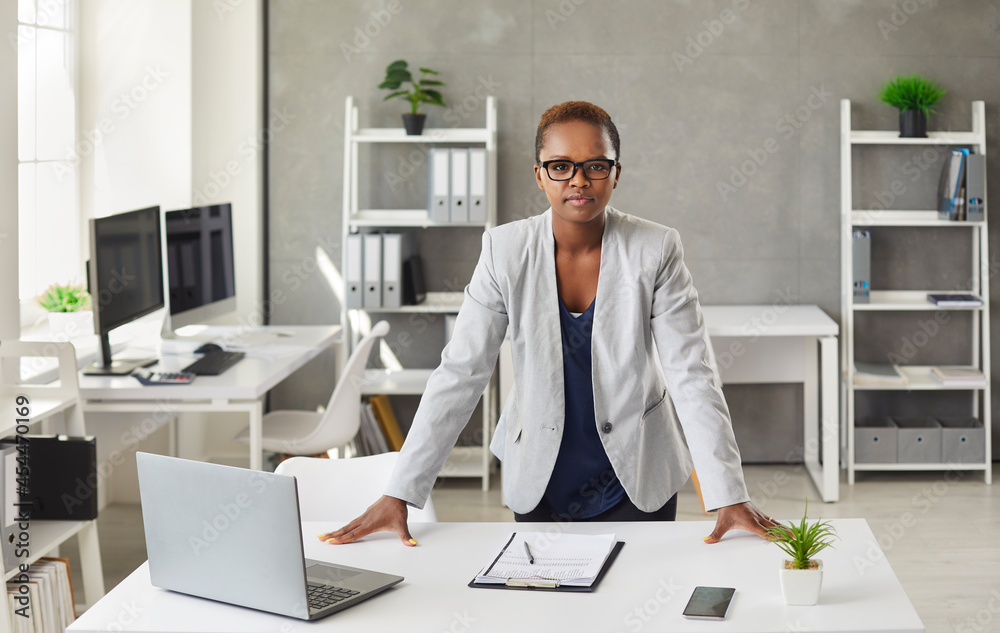 Portrait of a young african american businesswoman woman standing at her desk in the office. Woman with a short haircut and glasses is staring intently at the drain camera in the corporate office.