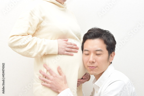 A young husband listens to fetal movements by listening to the large belly of a pregnant white-dressed young woman.
