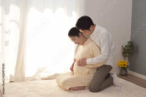 A young husband is touching the big belly of a pregnant white-dressed young woman from behind.