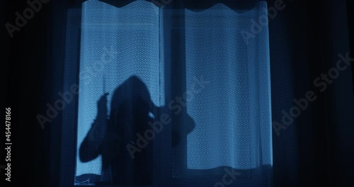 Killer is Looking through the Window at Night Holding a Knife Preparing to Break in the House in Silhouette
