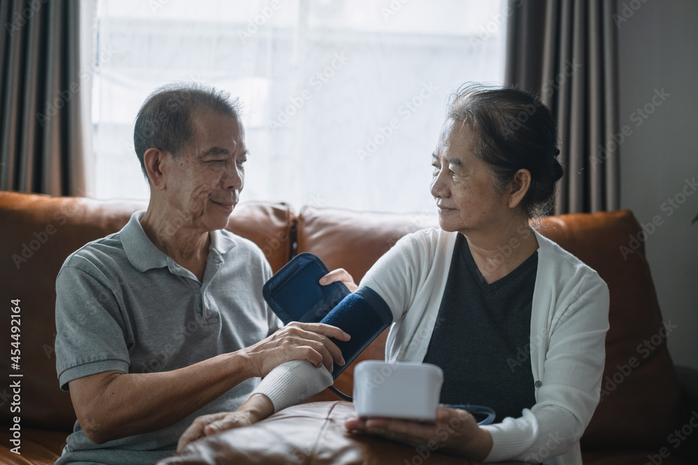 Asian senior couple checking blood pressure at home, a senior man is helping his wife wearing an arm cuff of the blood pressure gauge.
