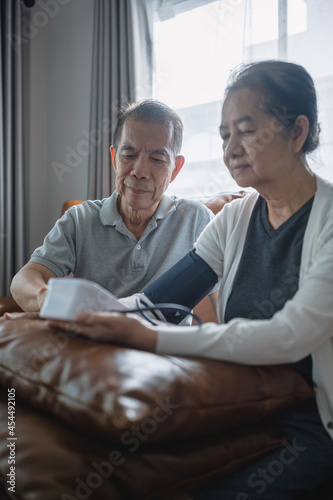 Asian senior couple checking blood pressure at home, a senior man is helping his wife wearing an arm cuff of the blood pressure gauge then press the start button.
