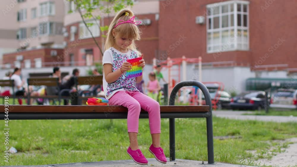 Joyful girl playing with pop it sensory anti-stress toy on playground bench. Child kid presses on colorful rainbow bubbles squishy. Stress, anxiety relief. Trendy game for stressed children and adults Joyful girl playing with pop it sensory anti-stress toy on playground bench. Child kid presses on colorful rainbow bubbles squishy. Stress, anxiety relief. Trendy game for stressed children and adults