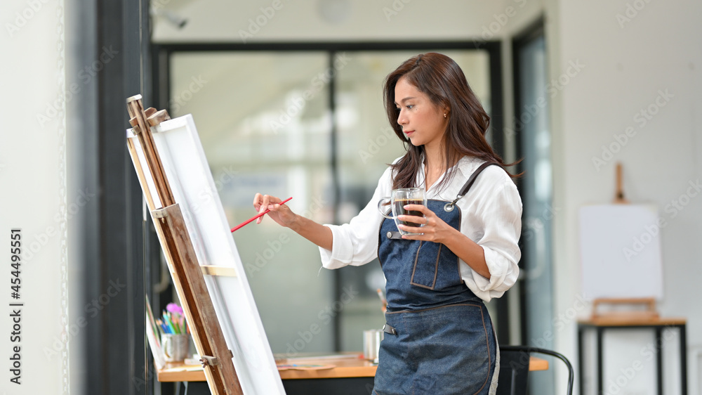 Obraz premium Hipster female painter paints on large canvas easel and holding coffee cup
