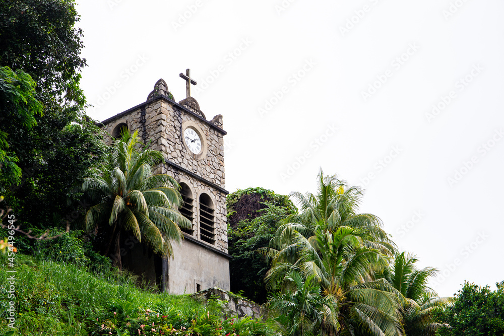 Stone Clock Tower (Tour de l'Horloge) on the hill next to the Immaculate Conception Cathedral, old building with clock and Holy Cross, Victoria, Mahe Island, Seychelles.