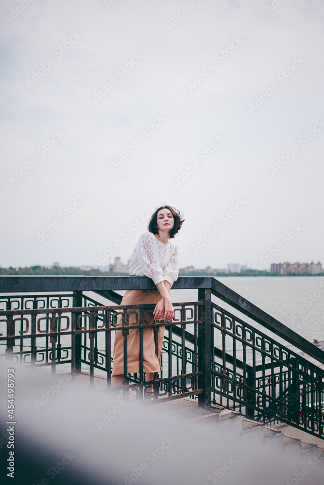 A happy girl on the river bank, standing on the bridge, smiling, a girl walks on the bridge by the river. Freedom. The wind is blowing the girl's hair.