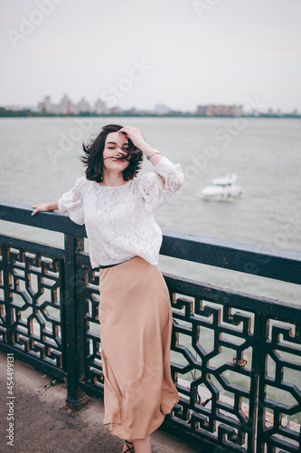 A happy girl on the river bank, standing on the bridge, smiling, a girl walks on the bridge by the river. Freedom. The wind is blowing the girl's hair. © Natalia Rzhevskaia
