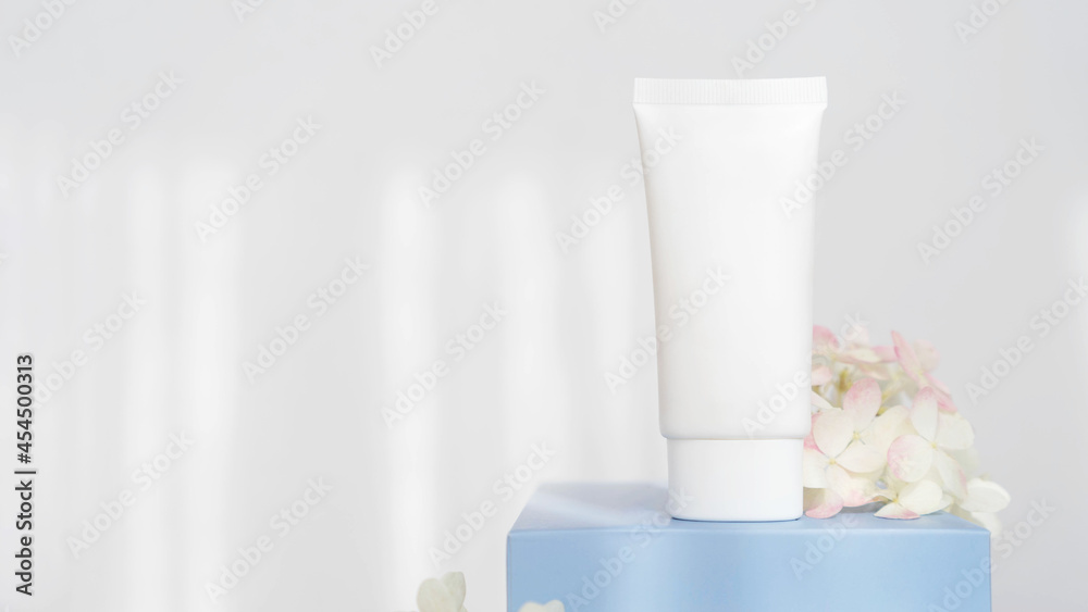 white cosmetic tube for cream on blue podium with flowers on white background with sunlight. Concept of gentle skin care. Copy space white cosmetic tube for cream on blue podium with flowers on white background with sunlight. Concept of gentle skin care. Copy space