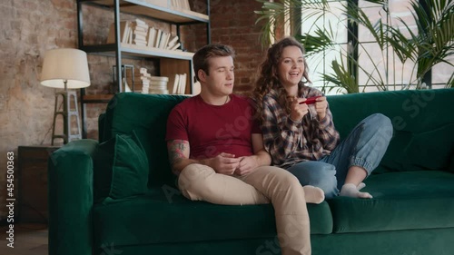 Young female sitting down on couch and grabbing controller from male gamer forcing man to take another gamepad and play videogame together on sofa at home