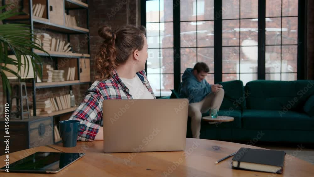 Pan right view of cheerful young woman asking boyfriend to join online conversation with friend while sitting at table and making video call on laptop at home Pan right view of cheerful young woman asking boyfriend to join online conversation with friend while sitting at table and making video call on laptop at home