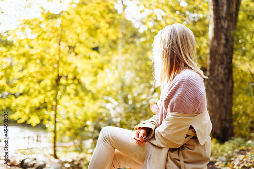 Lonely upset woman wearing cozy warm knitted sweater and coat sit in park at autumn sunny day photo