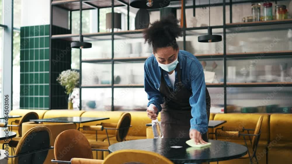 A african american cafe worker woman wearing protective medical mask cleaning the table inside the cafe A african american cafe worker woman wearing protective medical mask cleaning the table inside the cafe