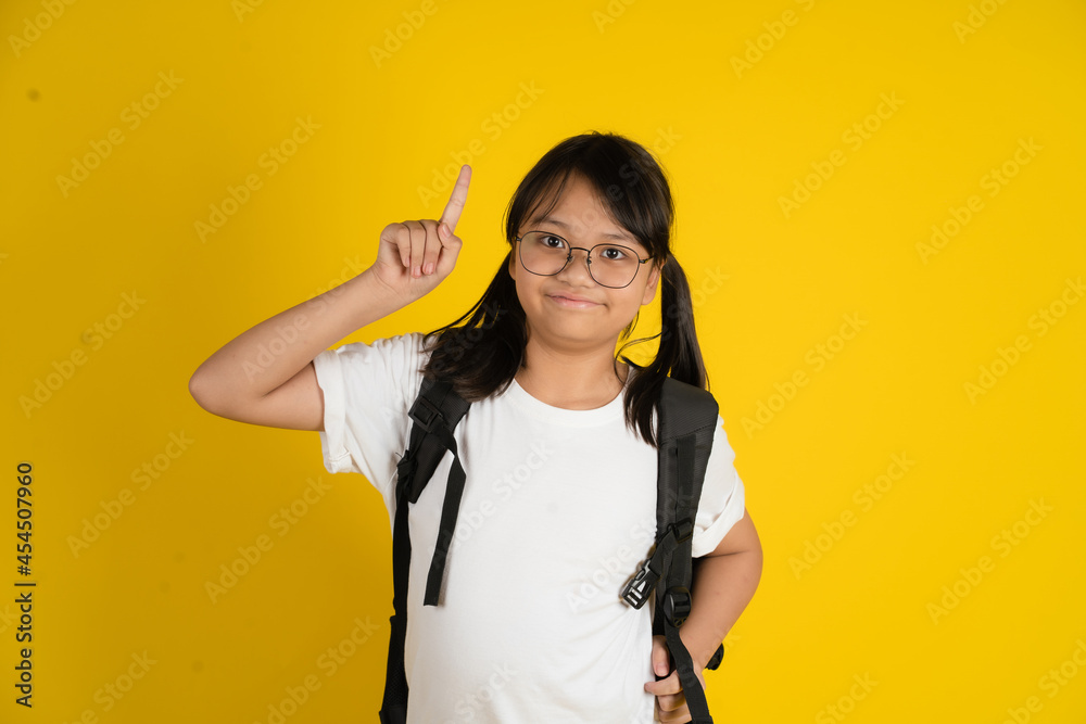 Little Asian girl wearing glasses and carrying a school bag with her finger pointing up, on a yellow background.
