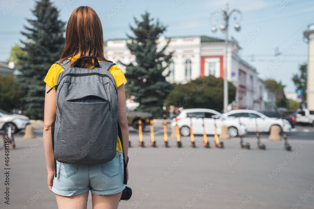 Woman travel in city with backpack, standing and looking on street with transport. Rear view and defocused background