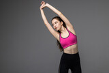 Young athletic fitness woman in sportswear stretching her arms and looking at camera while standing isolated over grey background. Healthy lifestyle and sports concept